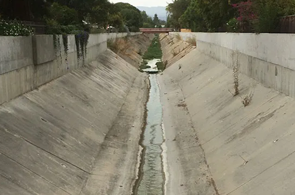 A trapezoidal concrete flood-control channel, bare gray walls rising on either side of a thin trickle of green water at the bottom