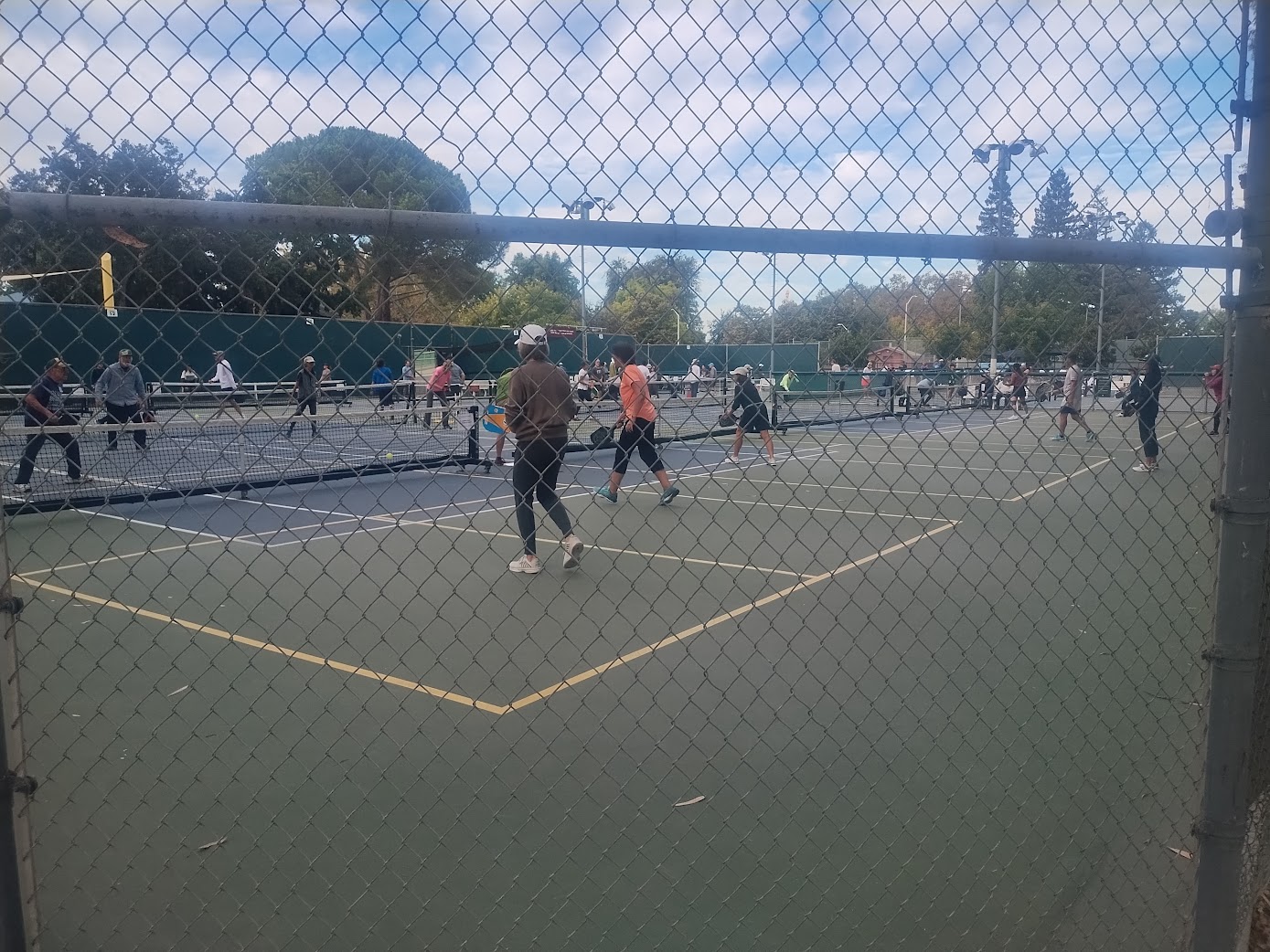 A crowded public pickleball court seen through a chain-link fence